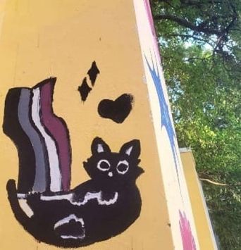 A small black cat painted on a yellow wall corner on Poovath Street, with a striped rainbow tail and a heart beside it.