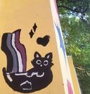 A small black cat painted on a yellow wall corner on Poovath Street, with a striped rainbow tail and a heart beside it.