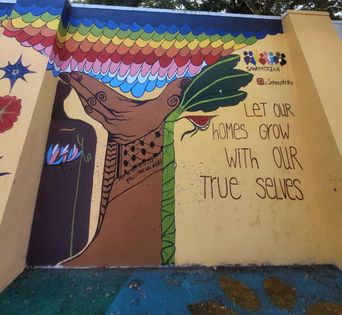 A wall mural on Poovath Street of a hand cradling a banana leaf and pomegranate under a scalloped rainbow, with the words ‘Let our homes grow with our true selves’.