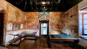A palace room with murals covering all four walls and a dark carved wooden ceiling, two low display tables on the polished floor and a doorway through to the next room.