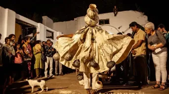 A performer in a wide gold costume with stacked pots balanced on their head, spinning at night in a courtyard as a crowd watches and a dog wanders past.