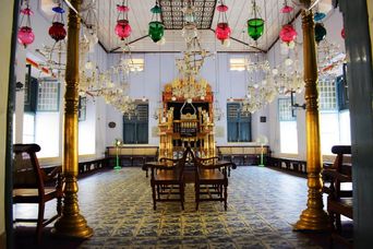 The synagogue’s blue-tiled interior seen between two gold pillars, colourful glass oil lamps hanging from the ceiling and the gilded ark at the far end.