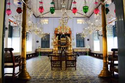 The synagogue’s blue-tiled interior seen between two gold pillars, colourful glass oil lamps hanging from the ceiling and the gilded ark at the far end.