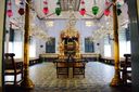 The synagogue’s blue-tiled interior seen between two gold pillars, colourful glass oil lamps hanging from the ceiling and the gilded ark at the far end.