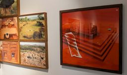 Gallery wall at Kara showing framed aerial photographs of an archaeological excavation beside a large red-toned artwork of stone steps and carved reliefs.