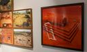 Gallery wall at Kara showing framed aerial photographs of an archaeological excavation beside a large red-toned artwork of stone steps and carved reliefs.