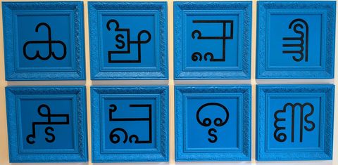 Eight blue-framed panels of black abstract line symbols mounted in a grid on a gallery wall at Muziris Contemporary.