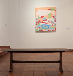 A wooden bench on a tiled floor facing a colourful abstract painting of houses on a gallery wall at Muziris Contemporary.