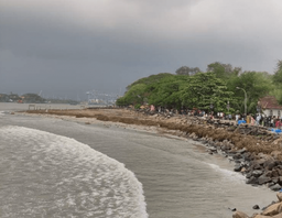 Rocky shoreline promenade with crowds walking under trees, port cranes across the water.