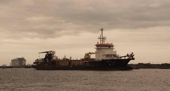 Industrial dredging ship anchored offshore under an overcast sky, a storage tank on the far shore.