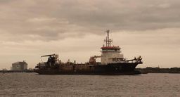Industrial dredging ship anchored offshore under an overcast sky, a storage tank on the far shore.