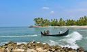 Fishermen in a black wooden boat riding a wave near a rocky breakwater, palm-lined beach behind.