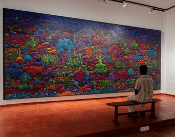 A man sits on a bench facing a large, vividly coloured mural of a forest full of figures and animals at Muziris Contemporary.