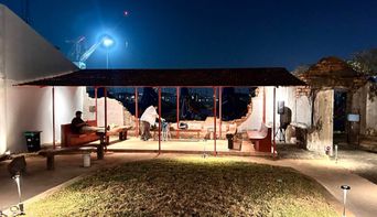 A lit open-sided pavilion in a courtyard at dusk, its tiled roof held on slim red posts, with a couple of people sitting inside and a floodlight above.