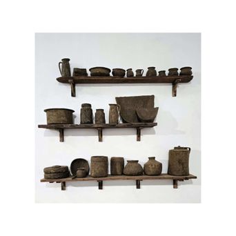 Three rough wooden shelves on a white wall, each lined with grey clay pots, jars, bowls and a grinding stone, all in the same dusty earth colour.