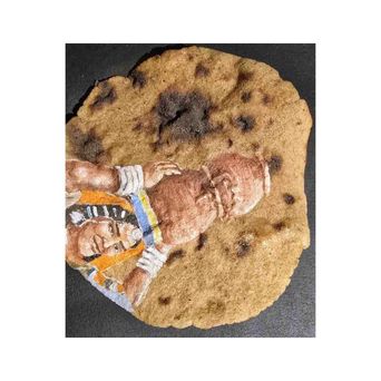 A blistered flatbread with a figure painted across it — a person in a striped shawl and beaded collar, lying curled on the browned surface.