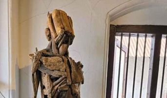 A standing figure built from lashed pieces of driftwood and scrap timber, placed against a cracked white wall beside a barred window.