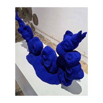 A sculpture on a white plinth: a cluster of faces and rounded forms modelled in vivid ultramarine blue, rising in a curve.
