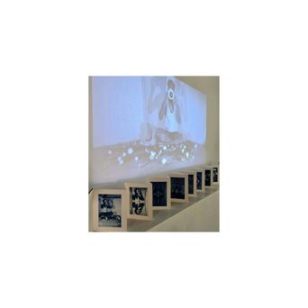 A row of small framed photographs standing along a low white ledge, a large dim projection of a seated figure filling the wall above them.