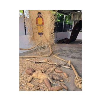 An installation in an open yard: a woven jute hanging painted with a figure in a yellow and red dress, split cane fanning out around it, and shaped pieces of wood laid out on the ground below.