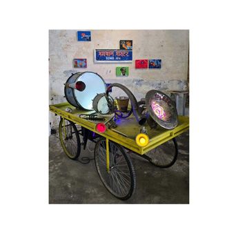 A yellow handcart loaded with brass band instruments — a big drum, a snare, a coiled horn and a wide bell lit from inside — parked against a peeling wall hung with small painted panels.