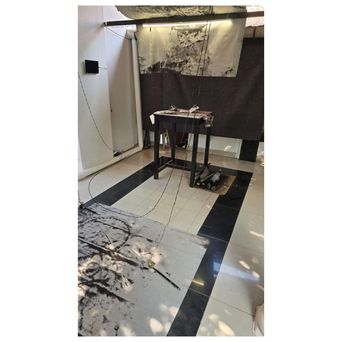 A small dark table rigged with wires in a bare room, a smudged black circle printed on the tiled floor and an ink-marked cloth hanging on the wall behind.