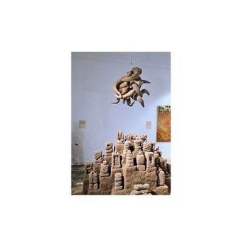A mound of small clay figures and tower shapes built up like a hillside village, with a knotted clay form hanging on a wire above it.