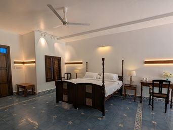 A bedroom with a dark wooden four-poster bed, blue patterned floor tiles, wall-mounted lights, a wooden desk and chair on the right, and a ceiling fan overhead.