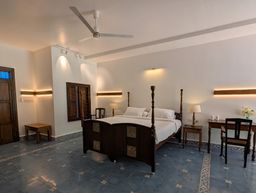A bedroom with a dark wooden four-poster bed, blue patterned floor tiles, wall-mounted lights, a wooden desk and chair on the right, and a ceiling fan overhead.