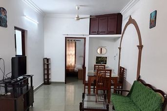 Living room with a flat-screen television on a wooden stand, a green upholstered wooden sofa, a glass-topped coffee table and a dining table with chairs beyond an arch.