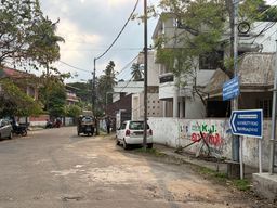 A paved road curves gently through a residential area lined with houses, trees, and utility poles with overhead wires. On the right, a white car is parked beside a low wall covered in colorful graffiti, next to a blue directional sign mounted on a pole. Further down the road, an auto-rickshaw and a motorcycle are visible, with a few people standing nearby. The sky is partly cloudy, and the scene is framed by lush greenery on both sides.