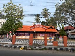 A temple building with a red tiled roof and a decorative arched sign above the entrance stands behind a metal fence supported by orange pillars. The building has pink and white walls with a patterned facade. Palm trees and other leafy trees rise behind the structure against a sky crisscrossed by overhead wires. A paved area with a black and white checkered pattern lies in front of the fence, and a road runs along the bottom edge of the scene.