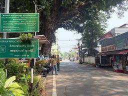 A green road sign stands on the left side of a paved street, with another green sign mounted above it. Several people stand near the signpost beside parked scooters and plants. A large tree with a thick trunk and dense foliage overhangs the road from the left. On the right, a row of buildings includes a shop with an open front and a red awning. Vehicles travel down the road into the distance under a bright sky.
