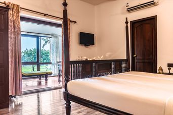 Guest room with a carved four-poster bed and wooden floor, its glass doors open onto a sit-out facing a lawn and coconut palms.