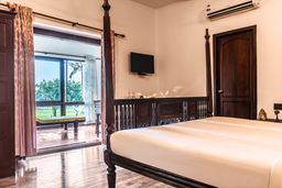 Guest room with a carved four-poster bed and wooden floor, its glass doors open onto a sit-out facing a lawn and coconut palms.