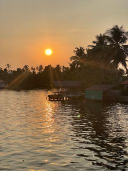 Sunset over a calm backwater, with palm trees along the far bank and a small wooden jetty and green-roofed shed on the water’s edge.