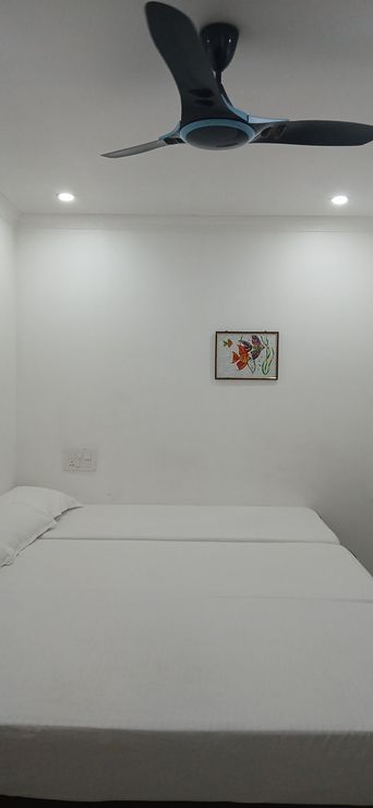 A white-walled room with two beds covered in white sheets, a dark ceiling fan overhead, a small framed picture on the wall, and recessed ceiling lights.