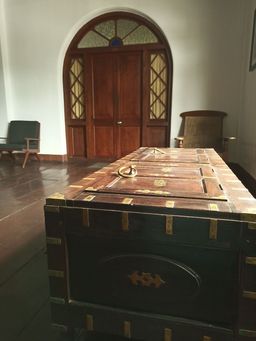 Large dark wooden chest with brass fittings and a brass ring handle on a dark wooden floor, in front of an arched wooden door with coloured glass panels and two armchairs.