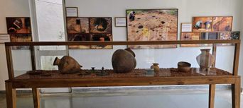 A long glass museum case holding large earthenware burial urns and small iron objects on a bed of red soil, with framed excavation photographs on the wall behind.