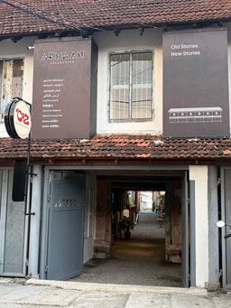 A two-story building with a tiled roof features two large brown panels on the upper facade, one on each side of a window. A circular sign hangs from a pole on the left side of the building. Below, an open grey gate leads into a covered passageway with a gravel floor, revealing a narrow path extending into the distance.