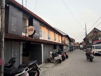 A narrow paved street lined with two-story buildings featuring tiled roofs and storefronts on the ground floor. Several motorcycles and scooters are parked along the left side, with one person riding a motorcycle away from the camera on the right side of the road. A white van is visible further down the street, and overhead utility wires run across the sky.
