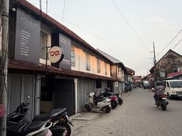 A narrow paved street lined with two-story buildings featuring tiled roofs and storefronts on the ground floor. Several motorcycles and scooters are parked along the left side, with one person riding a motorcycle away from the camera on the right side of the road. A white van is visible further down the street, and overhead utility wires run across the sky.
