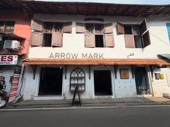 A two-story white building with a tiled roof overhang and wooden shutters on the upper windows. The ground floor has three large openings, with a freestanding sign in front of the central arched window. A small plaque is mounted on the wall to the right of the center opening. To the left, part of an adjacent building is visible with a sign and an air conditioning unit. A motorcycle is parked at the far left edge. The street runs along the front of the building.