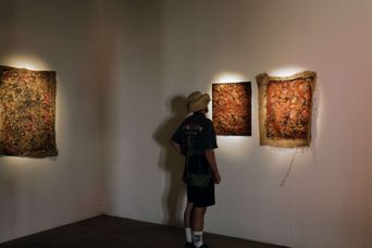 A visitor in a sun hat looks at large rust-orange textured textile artworks mounted on a white gallery wall at Aspinwall House.