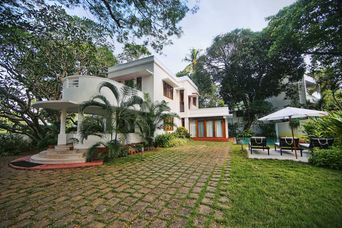 Two-storey white house with a curved balcony and stone steps, facing a cobbled drive, a lawn, tall trees, and a pool with loungers under a parasol.