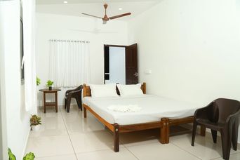 Bedroom with a wooden double bed under white bedding, a ceiling fan, white curtains at the window, a small round side table and brown plastic chairs on a tiled floor.
