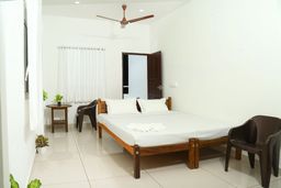 Bedroom with a wooden double bed under white bedding, a ceiling fan, white curtains at the window, a small round side table and brown plastic chairs on a tiled floor.