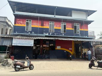A two-story building with a red, yellow, and blue painted facade stands on a dirt road. The upper floor has a covered balcony with a metal railing and several windows. Below, a corrugated metal awning extends over the ground floor, which features multiple entrances and a large painted section. A banner hangs on the left side of the ground floor. Several scooters are parked in front of the building, and a few people are visible near the entrances. To the left, part of an adjacent building and a low wall are visible, while to the right, a fence and some greenery can be seen.