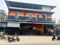 A two-story building with a red, yellow, and blue painted facade stands on a dirt road. The upper floor has a covered balcony with a metal railing and several windows. Below, a corrugated metal awning extends over the ground floor, which features multiple entrances and a large painted section. A banner hangs on the left side of the ground floor. Several scooters are parked in front of the building, and a few people are visible near the entrances. To the left, part of an adjacent building and a low wall are visible, while to the right, a fence and some greenery can be seen.
