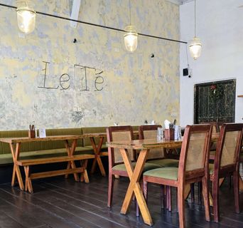 The cafe’s main room: bare plaster wall in patches of yellow and grey with "Le Té" spelled out in thin metal letters, glass pendant lamps, a long green bench behind wooden trestle tables, and cane-backed chairs on a dark wood floor.